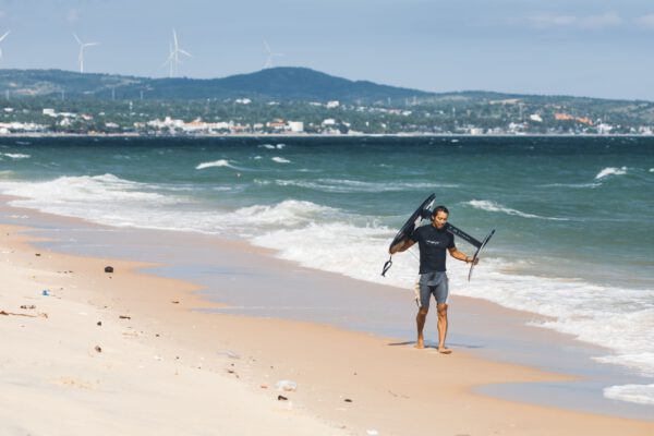A kitesurfer carries equipment along a sunny beach with wind turbines in the distance.